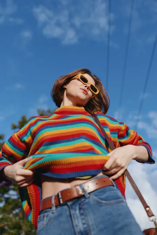 Woman, young woman in a colorful striped sweater and yellow sunglasses, pulling up sweater to show midriff in jeans outdoors under bright blue sky, playful confident travel style.