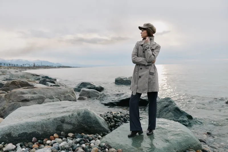 Woman in gray coat and hat stands on wet rocks by the sea looking sideways. Overcast sky and calm water create a serene coastal atmosphere with natural and fashion elements.