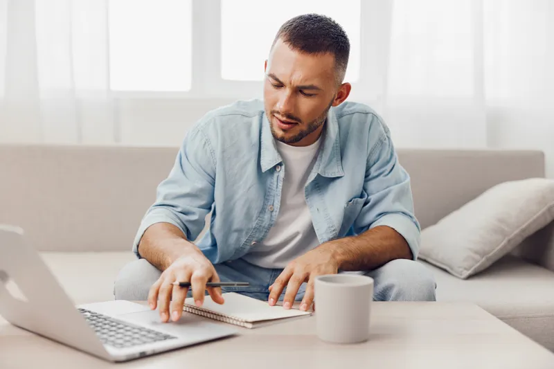 Thoughtful man working on a laptop while taking notes in a cozy home setting, showcasing productivity and creativity in a modern workspace.