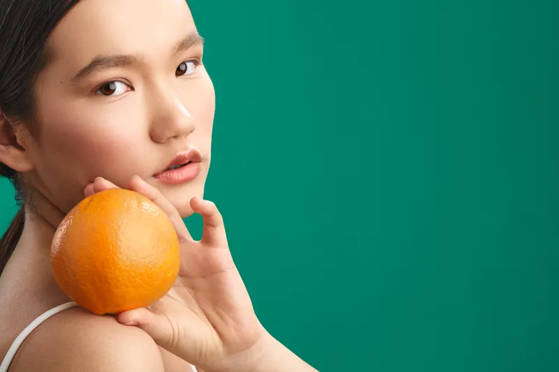 cheerful asian woman holds a bright orange near her shoulder in a clean studio setting, creating a fresh, vibrant mood with bold color contrast.