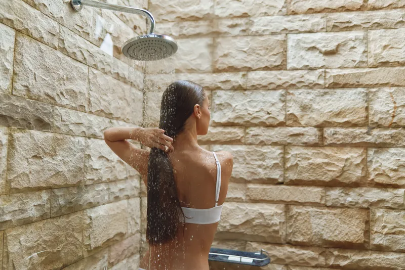 woman showering outdoors under rain shower head with wet long hair, natural stone wall background, peaceful moment of self-care and relaxation