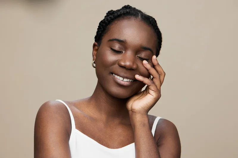 Young African woman with closed eyes smiling gently, wearing a simple white dress, exuding happiness against a soft beige background, perfect for wellness and beauty concepts