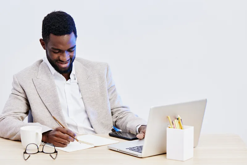 Smiling young African American man wearing beige blazer working at desk with laptop in bright modern office, using pen and notebook, concept of professionalism and productivity, workspace environment,