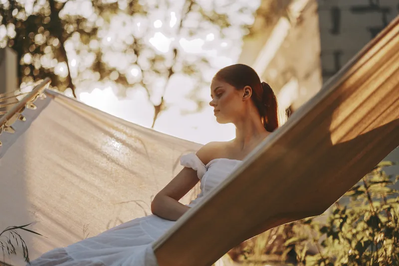 Young woman relaxing in a hammock outdoors, wearing a white dress, exuding tranquility and peace amidst a bright, sunny backdrop with natural greenery