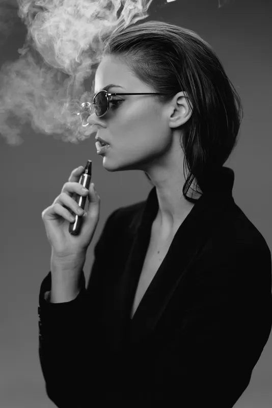 Black and white portrait of a woman vaping with sunglasses, stylish hair, and a confident expression in a studio setting.