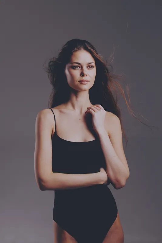 Young woman in black bodysuit posing with calm expression in studio with soft lighting and neutral background