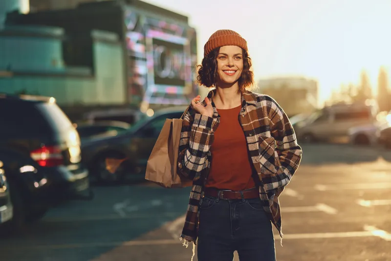 Woman shopping with paper bag in parking lot, smile glowing in warm sunlight during golden hour; candid lifestyle portrait highlighting authenticity, mindful living and emotional storytelling.