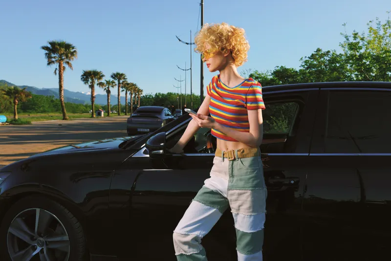 Woman lifestyle shot with retro film color, standing beside a sleek car on a sunny road, palm trees in the background, striped top and jeans, casual candid street mood