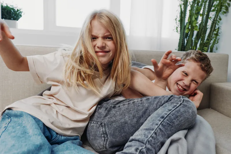 Playful siblings having fun on couch, laughing and enjoying energetic moment in cozy living room with natural light and casual clothes.