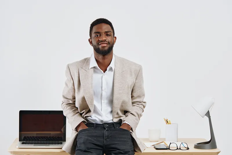 Confident young man with stylish blazer and white shirt standing in modern office with laptop and glasses; professional, relaxed, and approachable vibe. People lifestyle concept