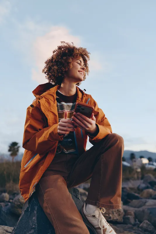 Woman lifestyle outdoor, a woman in orange jacket with curly hair smiles while holding a smartphone on the beach, casual communication, bright sky for travel lifestyle moment