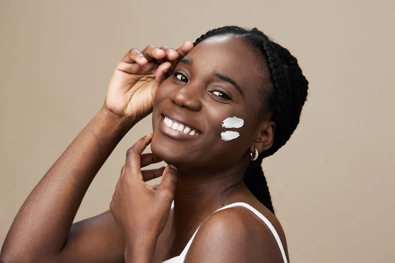 Smiling Black woman applying skincare with a neutral background, showcasing beauty, confidence, and self care in a minimalist style