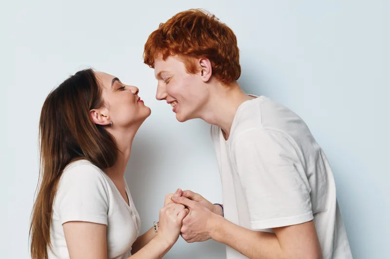 Young couple holding hands and leaning toward each other with smiles in casual white tops on plain background. Positive connection, love, relationship, happiness and affection moment.