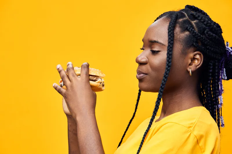 A young Black woman enjoying a delicious burger against a vibrant yellow background, showcasing joy and satisfaction in her casual yellow outfit
