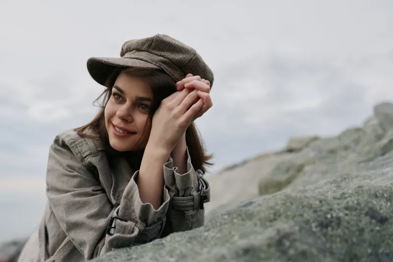 Smiling young woman in vintage hat and coat leans on rock outdoors with cloudy sky in background. Concept of autumn fashion, nature, and relaxed lifestyle in soft natural light.