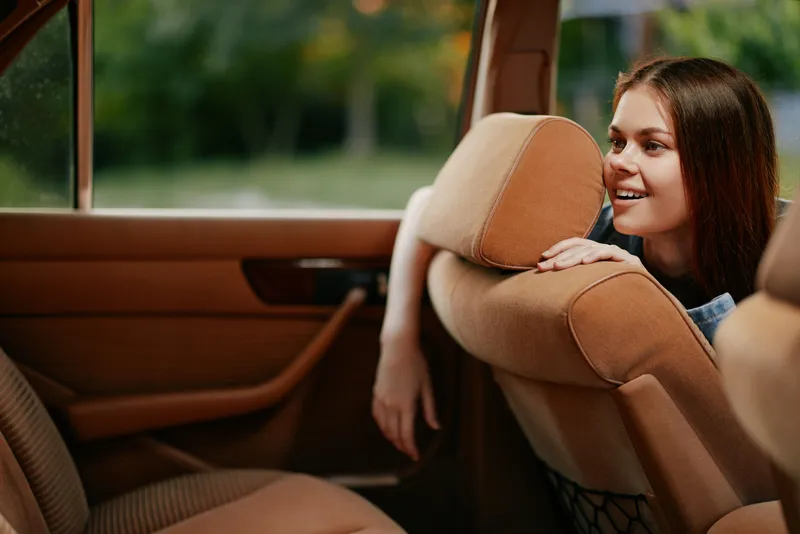 A cheerful young woman in casual attire smiling from the back seat of a car, showcasing an inviting atmosphere, perfect for travel and adventure themes.