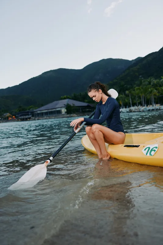 A solo paddler sits in a yellow kayak, paddling along a tranquil shore under a clear sky, with distant mountains and blue water, conveying focus, balance, and outdoor adventure.