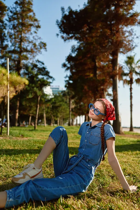 young woman relaxing on grass wearing blue jeans and sunglasses, red headscarf, sunny day in park with trees and clear sky visible