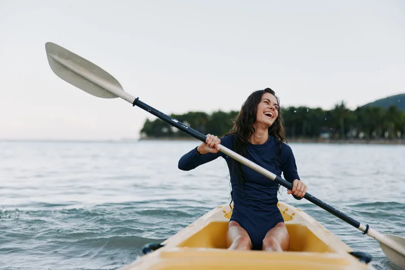 Joyful kayaker paddles across calm water in a bright yellow kayak, laughing with delight as the oar slices through gentle waves under a clear sky.