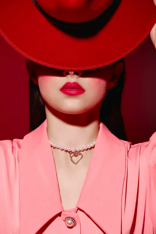 Stylish woman wearing bright red hat partially covers face emphasizing glänzende pink lips pearl necklace and pink outfit. Creative beauty fashion editorial with bold colors concept.