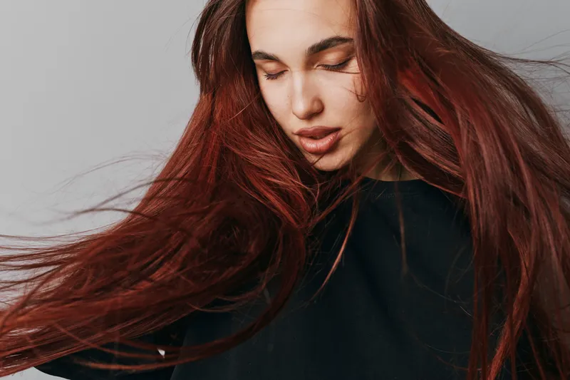 Young woman with long red hair and closed eyes wearing black sweater posing against gray background. Hair movement and natural beauty with peaceful expression in studio portrait.