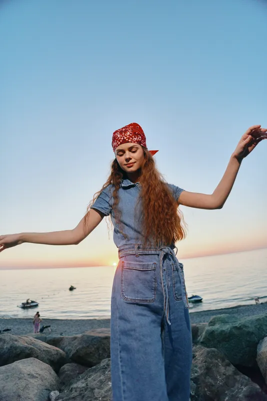 young woman dancing at sunset wearing denim dress and red bandana near rocky beach and calm sea water