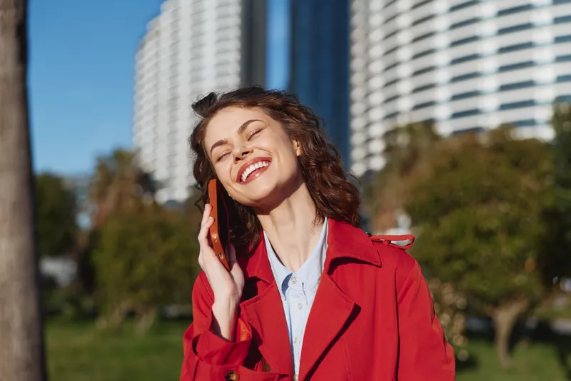 Young brunette woman with curly hair smiles joyfully while talking on a smartphone outdoors near modern buildings on a sunny day.