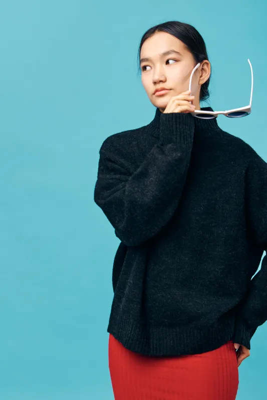 Young woman wearing black sweater holding sunglasses with hand looking sideways on blue background. Portrait of thoughtful female model with casual style for fashion or lifestyle concept.