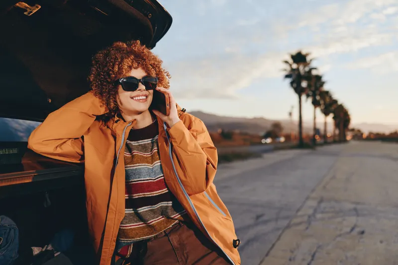Woman lifestyle outdoors, smiling near car at sunset, casual jacket, striped sweater, sunglasses on, relaxed travel pose by street, curly hair, cheerful smile, warm evening light