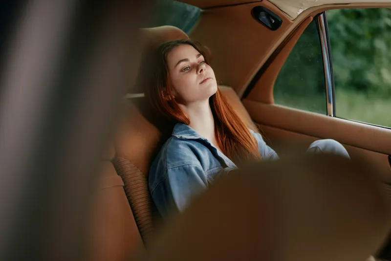 Thoughtful young woman with red hair resting in a vintage car, wearing a denim jacket, reflecting on life in nature, evoking calm emotions.