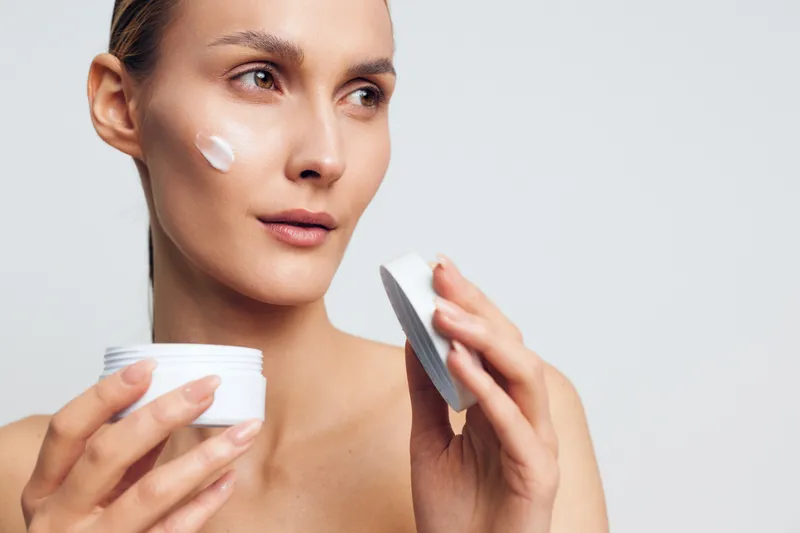 Woman applying face cream, skincare self care wellness close up, studio lighting, she smooths white cream on cheek with a spatula, holding a jar, beauty routine for healthy skin