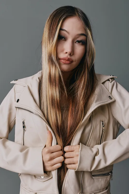 Young woman with long hair wearing beige jacket posing in studio with neutral background. Female model looking at camera with confident expression and casual fashion style.
