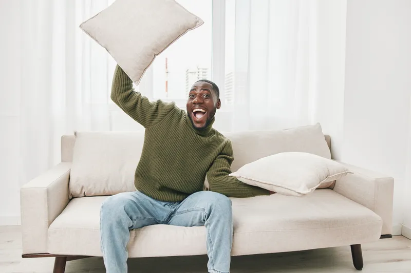 Joyful man with pillows on the couch in a bright living room, expressing happiness and playfulness through home comfort and relaxation
