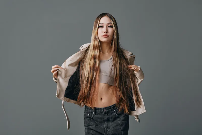 Young woman with long hair wearing crop top and open jacket posing confidently against gray background in casual fashion style for portrait or lifestyle concept.