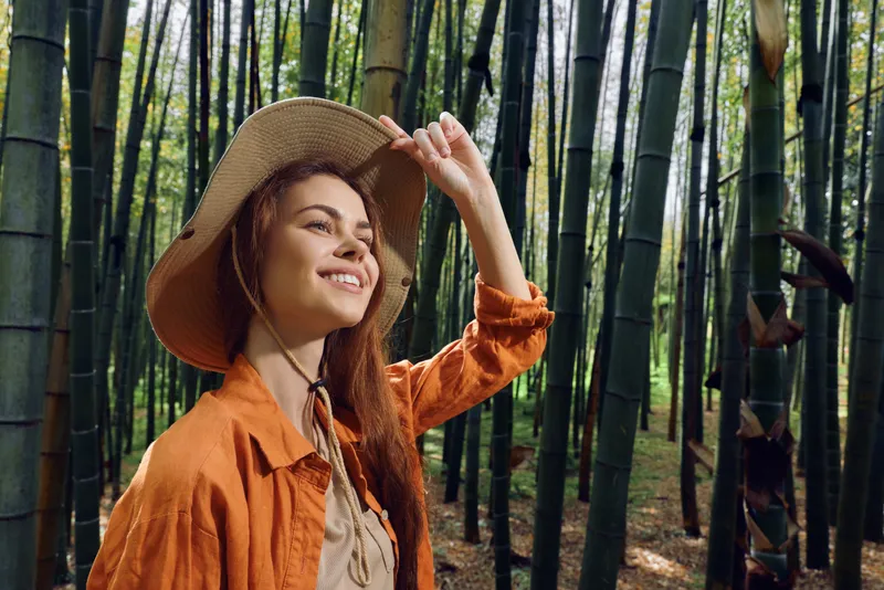 Woman bamboo forest hat smile nature portrait of a young traveler wearing a straw hat, looking up and enjoying sunlight among tall green stalks for a serene outdoor experience.