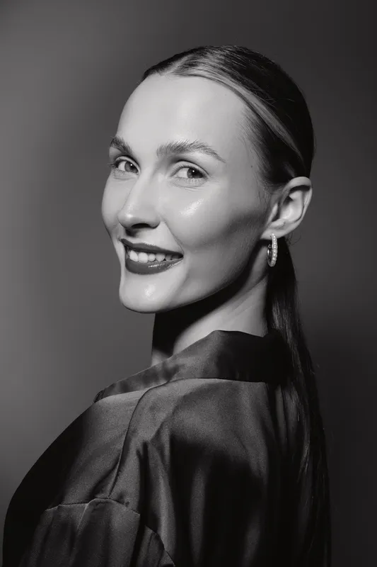 Portrait of a smiling Caucasian woman with light skin and slicked-back hair wearing hoop earrings and a satin blouse. Studio lighting highlights her clear complexion and confident gaze.