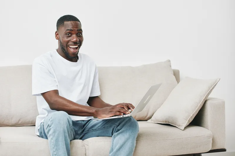 Young African man working on a laptop, smiling with joy, casual attire, sitting on a light sofa in a bright, modern room, conveying happiness and productivity
