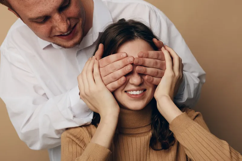 Smiling man covers eyes of happy woman in brown sweater with his hands. Intimate moment of surprise and affection in cozy indoor setting. Concept of love, trust, and relationship.