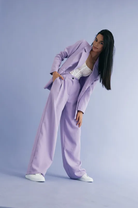woman in lilac suit standing on light purple background with white sneakers and lace top, stylish fashion look with long black hair and casual pose