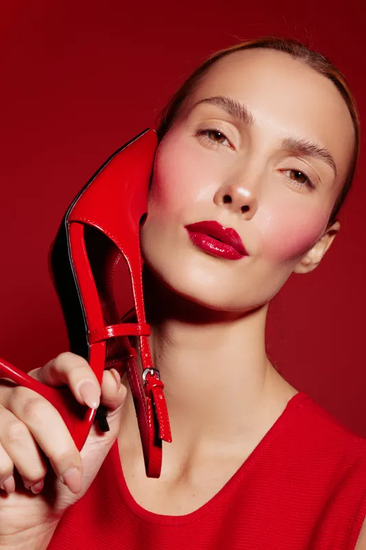 Close-up of a woman with glossy red lips and blush holding a red high heel shoe near her face. Red background and clothing create a bold fashion and beauty concept with strong color contrast.