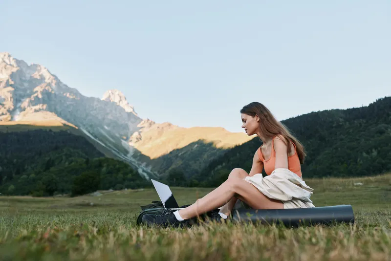 A focused young woman sits outdoors on a foam roller with a laptop, surrounded by mountains and a green meadow, creating a calm, productive outdoor workspace.