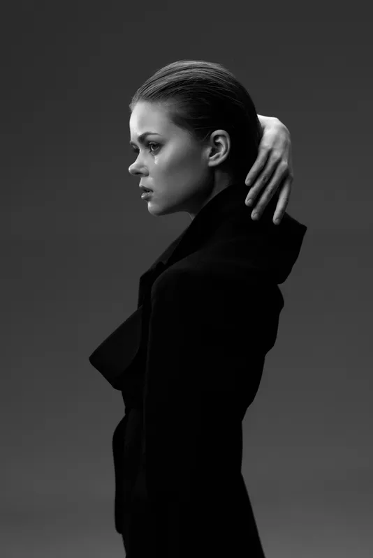 Black and white portrait of a serious woman in a dark coat with slicked-back hair and pensive expression.