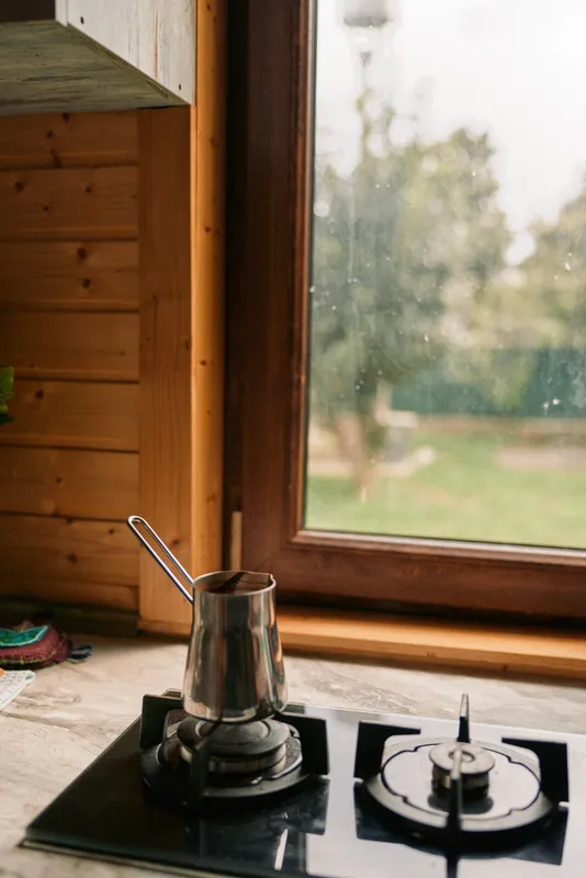 Calm kitchen scene shows a gas stove with a metal pot and kettle on the burner, framed by warm wood panels and a bright window, capturing a cozy home atmosphere.
