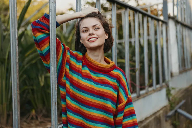 Woman smiling portrait in a colorful rainbow sweater, casual leaning pose by a greenhouse glass wall with plants, natural light and relaxed expression for lifestyle fashion.
