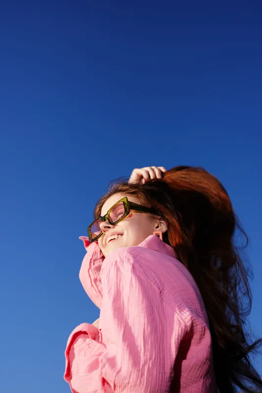 Happy young woman with long red hair wearing a pink shirt and stylish sunglasses against a bright blue sky, capturing a joyful and carefree essence of summer days