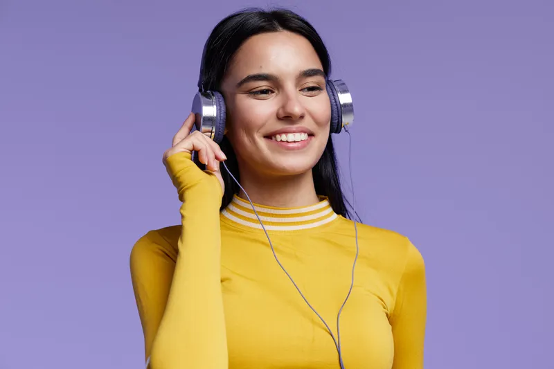 Sport fitness woman enjoying music with headphones, wearing a bright yellow top, smiling confidently against a purple background in a casual style.