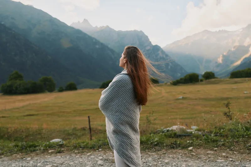 Young woman wrapped in a gray shawl stands in a windswept meadow, looking toward distant mountains, conveying calm, wonder, and quiet connection with nature