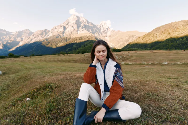 Woman portrait sitting in meadow with mountains in background, wearing shearling jacket and rubber boots, autumn outdoor scene, peaceful nature, relaxed young traveler.