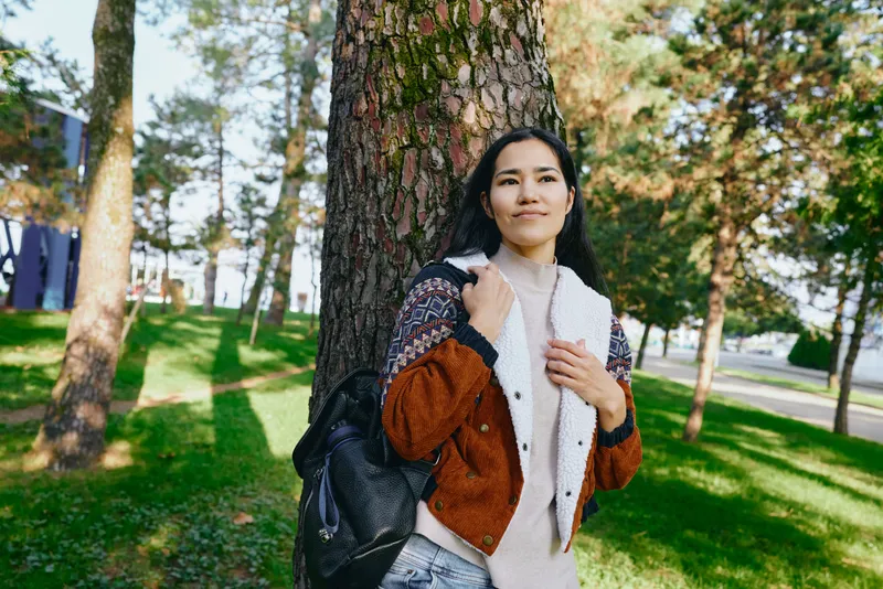 Woman with backpack leaning against a tree in a sunlit park, wearing a layered jacket and scarf, enjoying a calm outdoor moment and fresh autumn air, content and relaxed