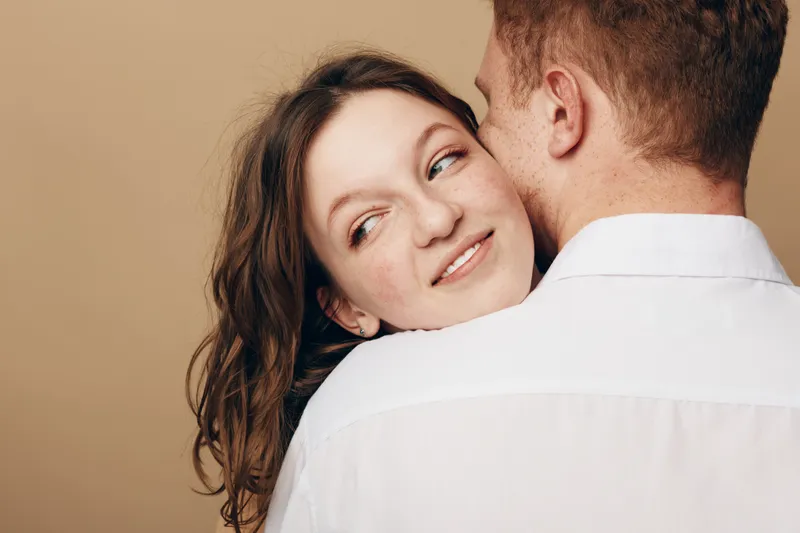 Smiling young woman with long brown hair embraces man in white shirt, looking over his shoulder with soft expression. Intimate moment, love, affection, and connection between couple in warm studio.